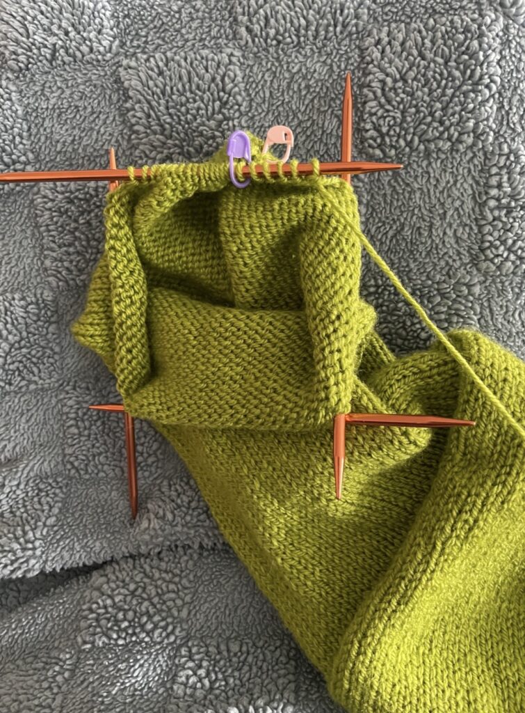 Four double pointed knitting needles used to knit the sleeve of a green sweater.