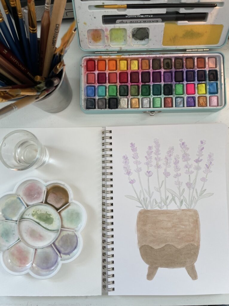 Watercolor workbook with painted picture of a flower pot holding tall little purple flowers. Around the book is a tray of watercolors, a cup of brushes, a glass of water, and a dish for mixing colors.