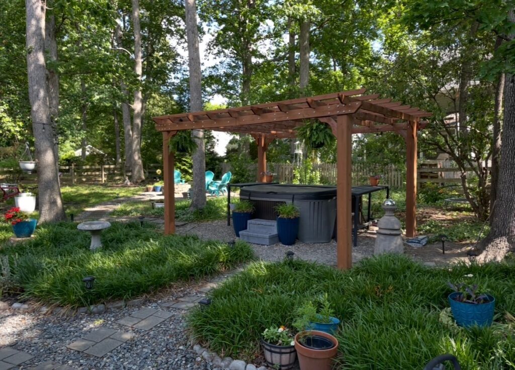 Wooden pergola over a hot tub in a backyard with gravel paths, green plants, and trees