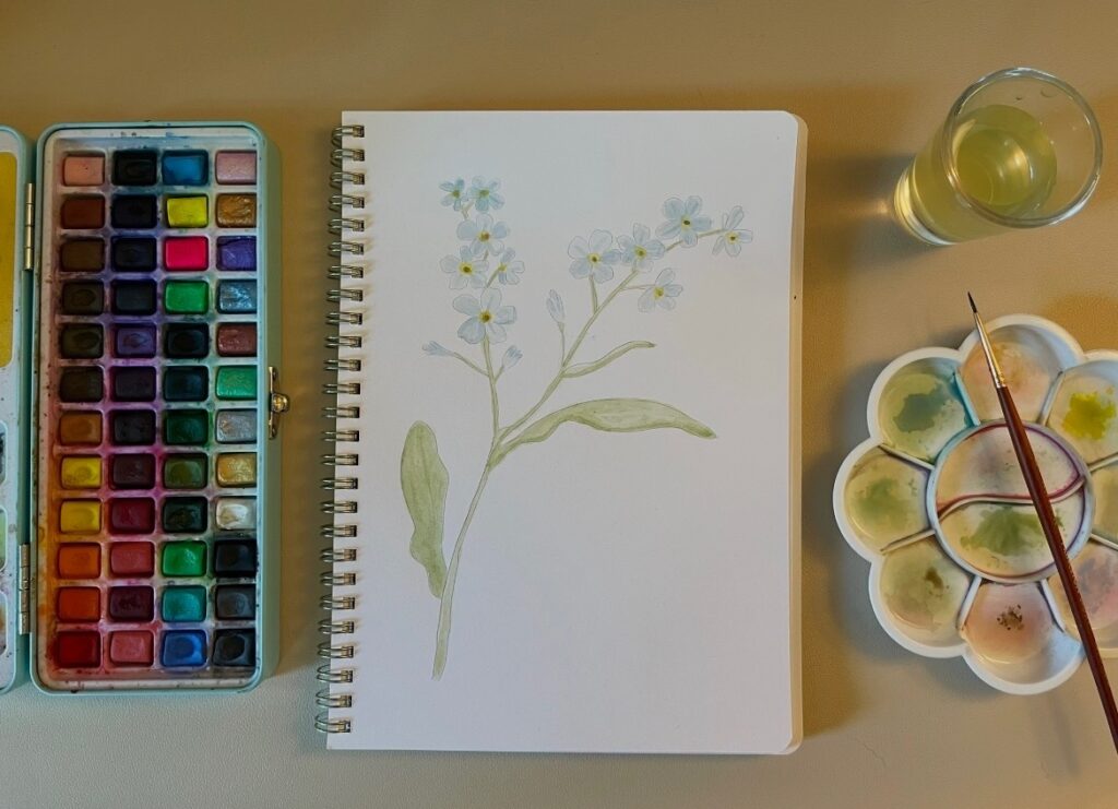 Watercolor of blue flowers with watercolor paint, a paint tray, a glass of water, and a paint brush.
