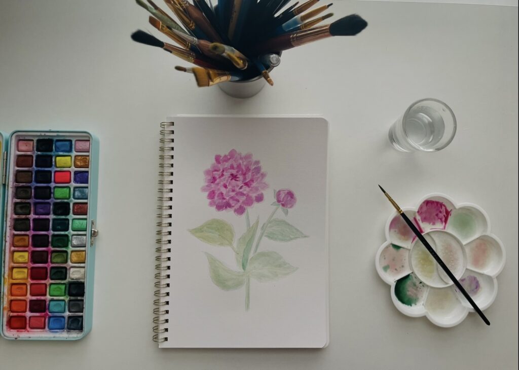 watercolor painting of a pink flower with paintbrushes, watercolors, and a a surrounding it.