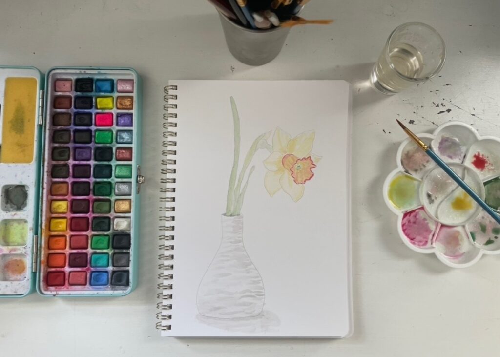 Watercolor painting of a daffodil with palate, paints, and brushes surrounding  it
