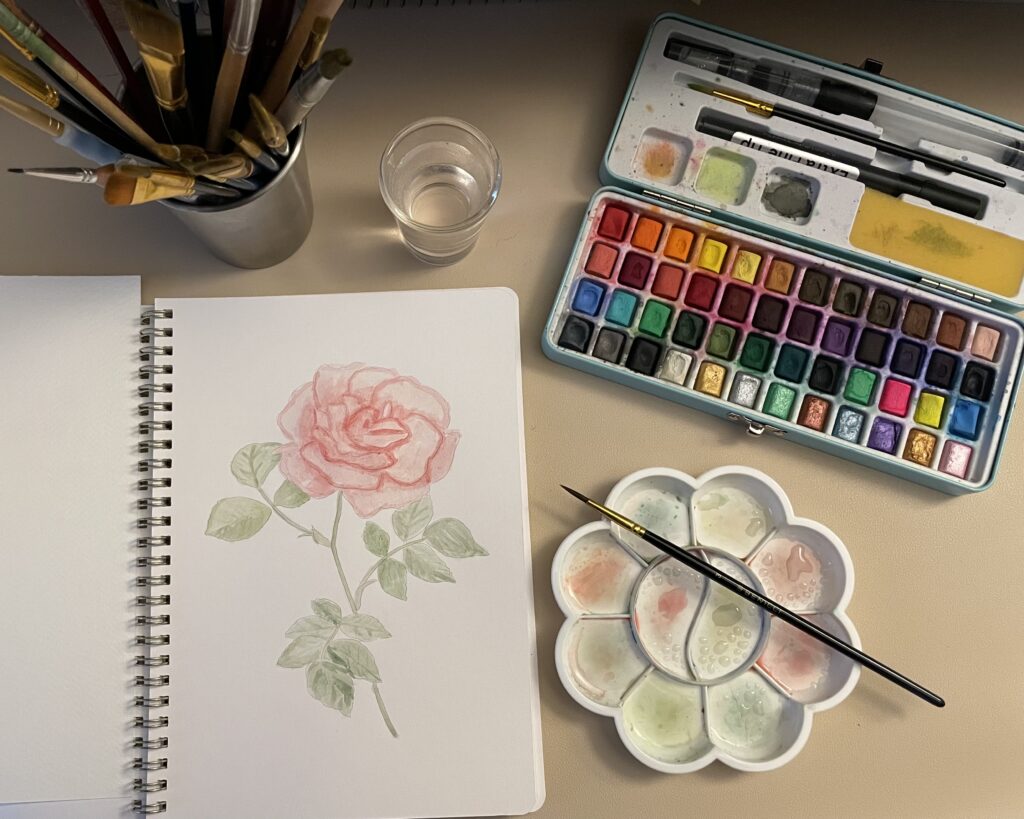 Watercolor workbook with painting of a pink flower. Around the workbook is a palate for the paint, paint, paint brushes, and a small glass of water.