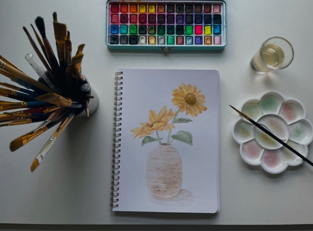 Watercolor painting of orange and yellow flowers. Around the painting are brushes, water, paints, and a mixing tray.